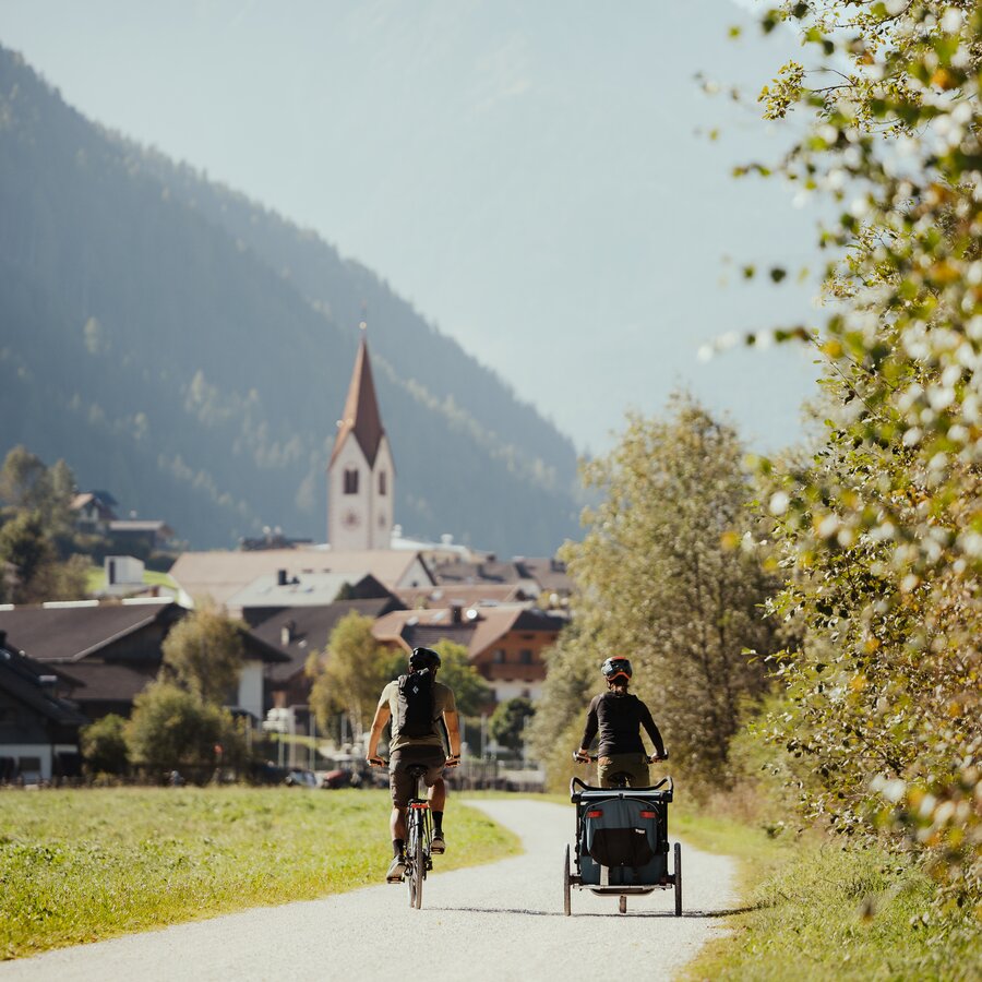 Zwei Radfahrer mit Kinderanhänger fahren auf einem Weg in Richtung Dorf Oberrasen mit Kirche und Bergen im Hintergrund. | © IDM Südtirol - Alex Molling