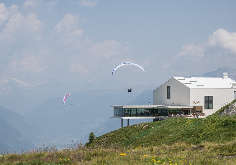 Zwei Paragleiter fliegen vor einem modernen weißen Gebäude mit Panoramaterrasse an einem Berghang. | © Harald Wisthaler