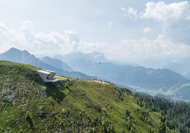Blick auf ein modernes Gebäude am Hang mit mehreren Gleitschirmen am Himmel über grüner Landschaft. | © Harald Wisthaler