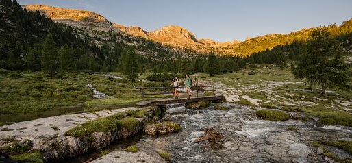 Eine Holzbrücke führt über ein flaches, felsiges Wasserbett mit Blick auf ein rötlich beleuchtetes Bergmassiv. | © Harald Wisthaler